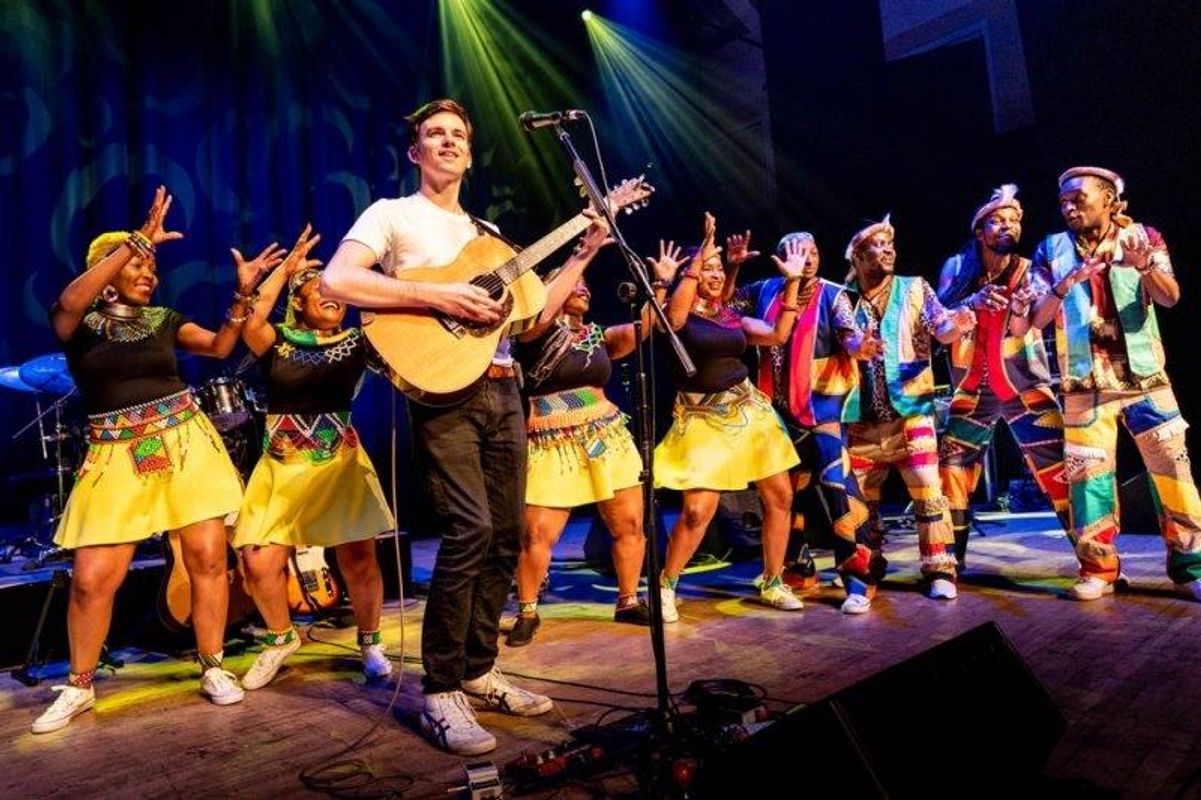 The South African Cultural Choir are singing on stage with Paul Simon. They are all wearing bright and vibrant African clothing. Paul is playing a guitar