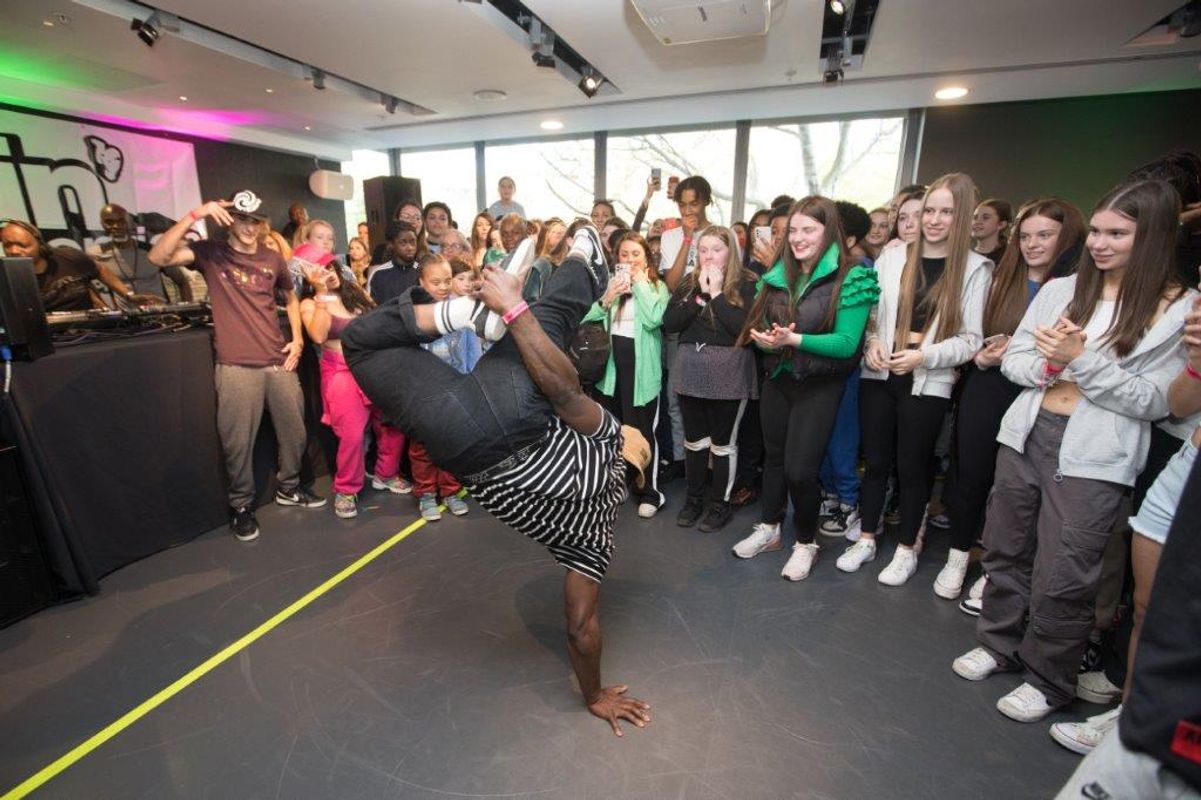 A person does a breakdance move balancing on one hand with a  group of young people surrounding in a semi circle.