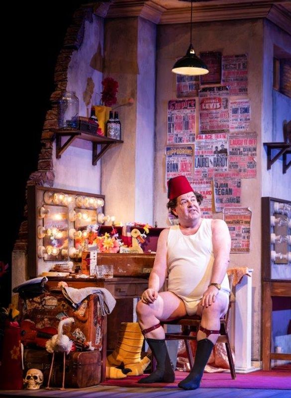 A man in a comedic outfit, wearing a fez and undershirt, sits onstage with an exaggerated expression. The room is cluttered and lively, featuring a vintage vanity with lights, posters, and props. The atmosphere is theatrical and whimsical, evoking humor and nostalgia.