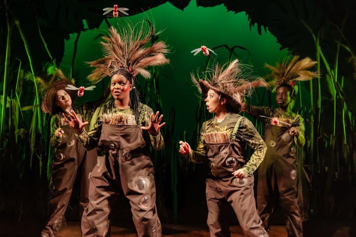 Four people stand on stage in brown dungarees and feathered hats.