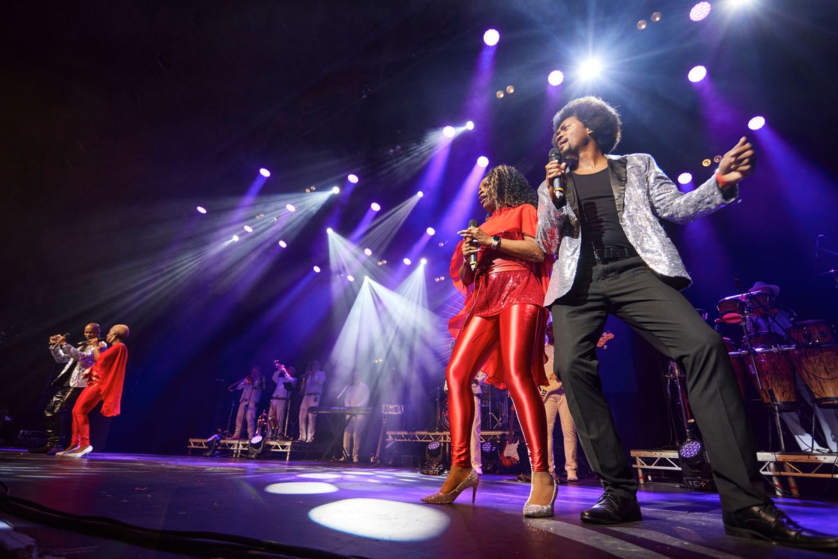 Multiple cast members stand on stage singing and dancing in pairs. The men wear a silver sequinned blazer and women  a red sequinned tunic, trousers and cape.