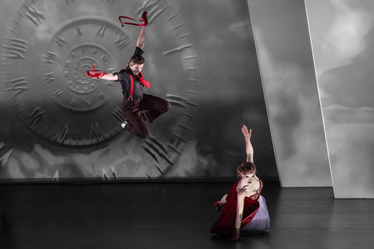 A dancer dressed in black and red leaps over another dancer.