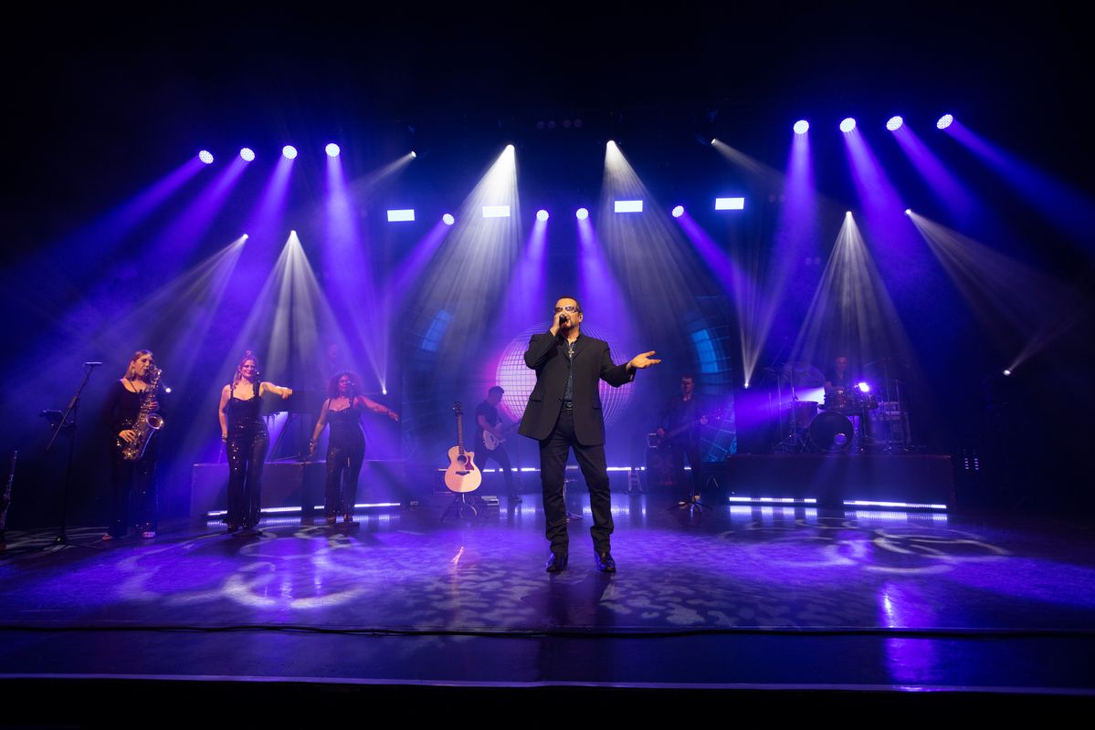 A man performing on stage to an audience. There are colourful lights. Behind him are backup singers and a band playing