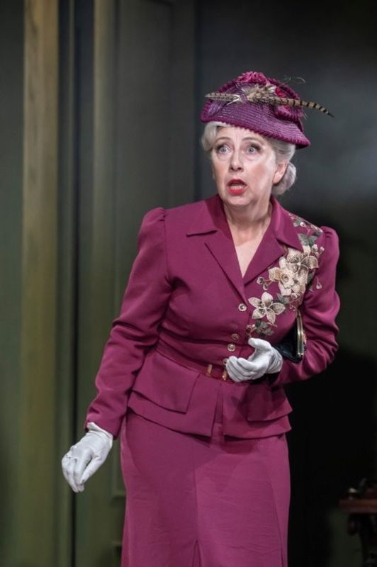 A woman wearing a magenta outfit adorned with floral embroidery and a matching hat decorated with a feather. She has a shocked look on her face.