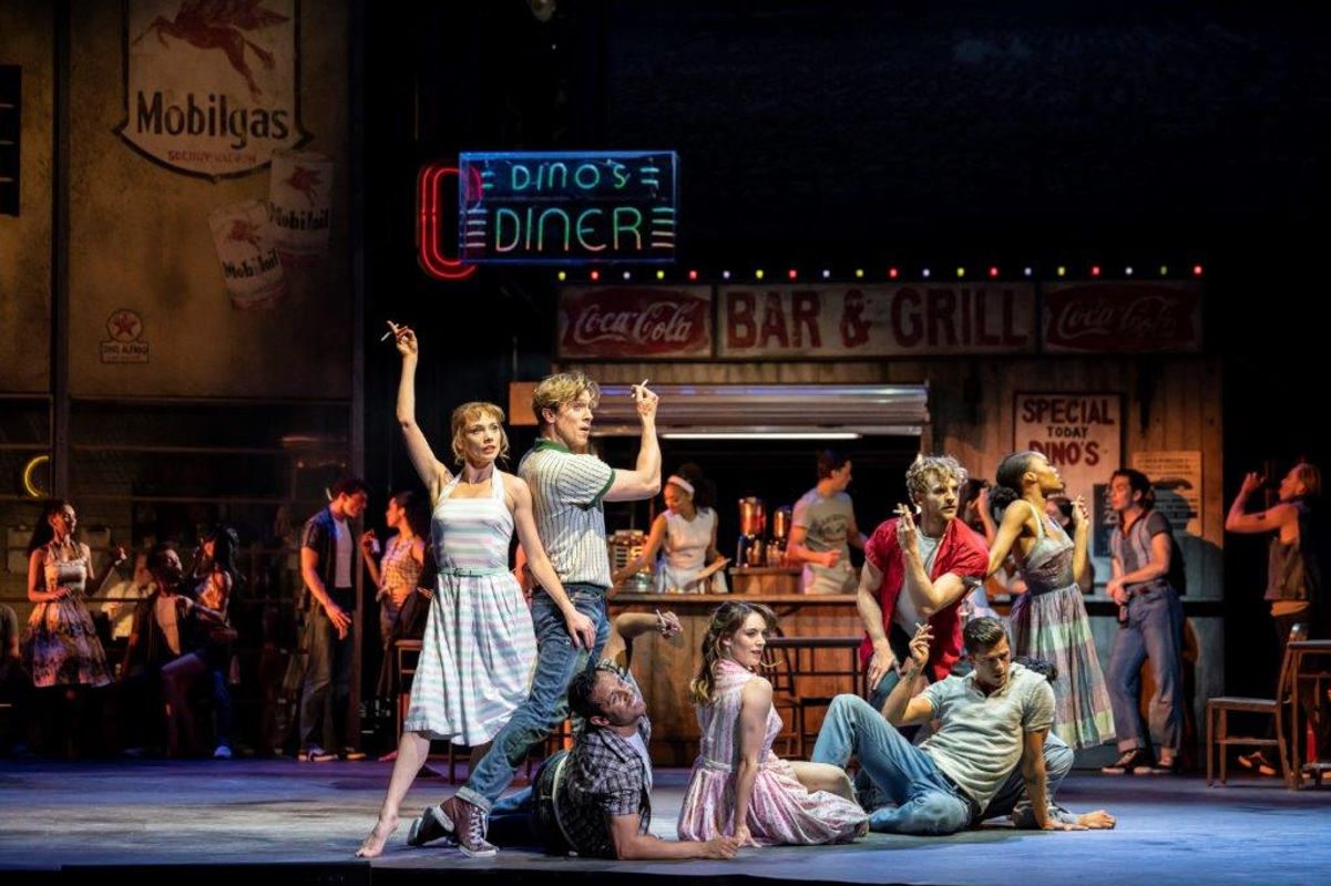 A lively 1950s diner scene with dancers in colorful retro attire striking energetic poses. Neon signs and a bar backdrop create a vibrant, nostalgic vibe.
