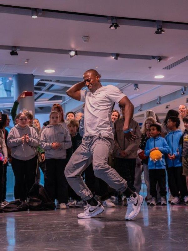 A person wearing a grey top and jeans bends both knees inwards and raises their right arm behind their head whilst being watched by a group of young people.
