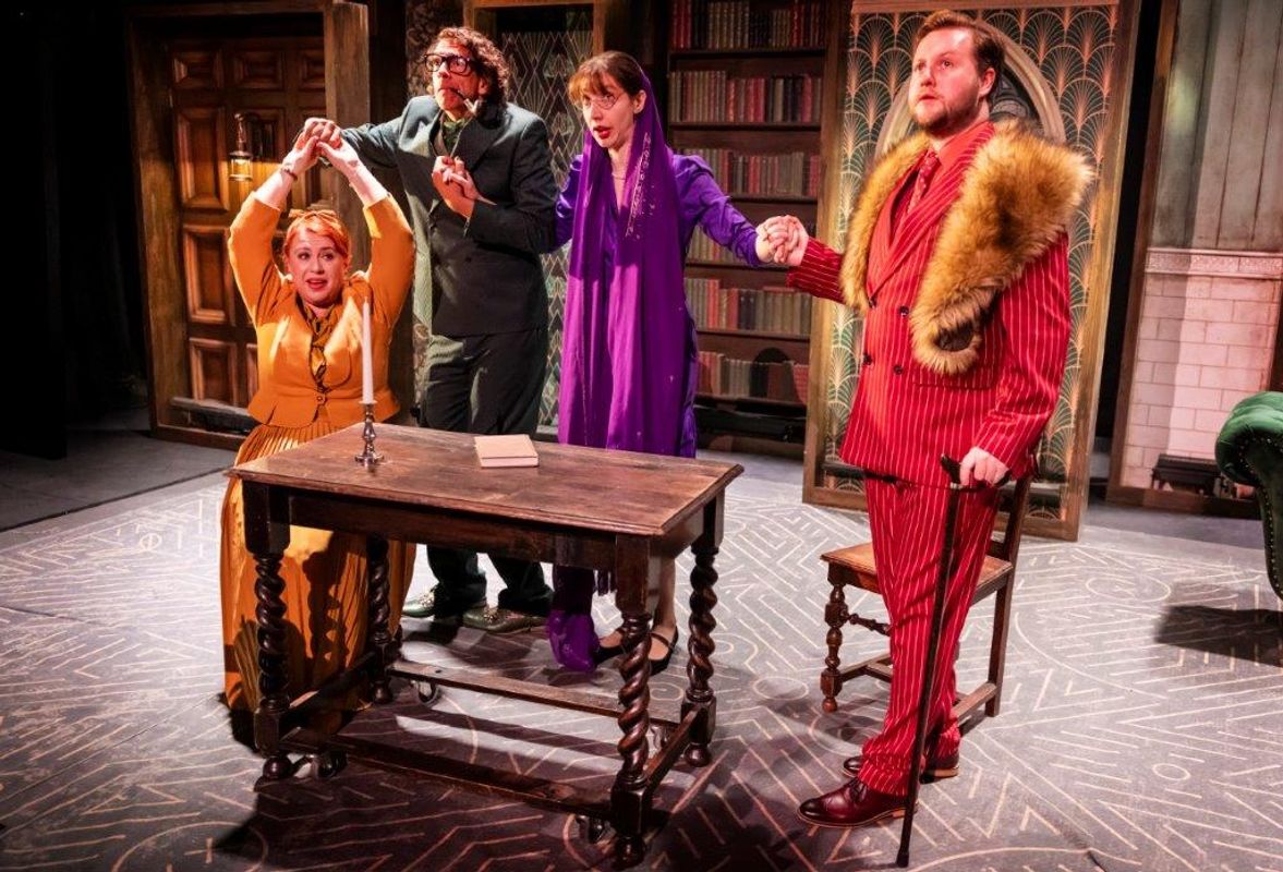 A group of four theatrical performers stands dramatically around a small wooden table. They wear colorful vintage costumes, exuding suspense and mystery.
