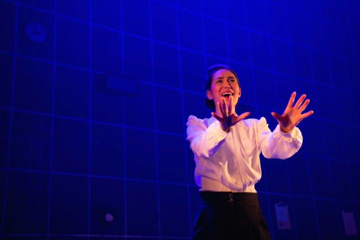 An actor in a white shirt performs on stage under dramatic blue lighting, with an expressive, intense gesture and facial expression, against a grid-patterned backdrop.