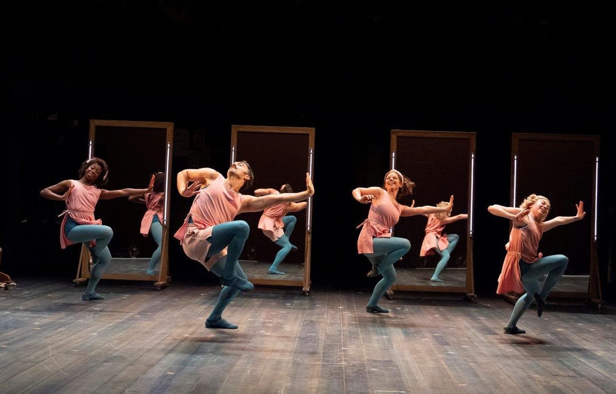 Dancers are performing with mirrors behind them. They are all wearing blue tights and pink dresses