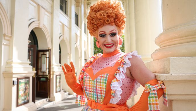 An actor playing the Dame waves at the camera wearing a bright orange wig and a chequered and polka dot dress.