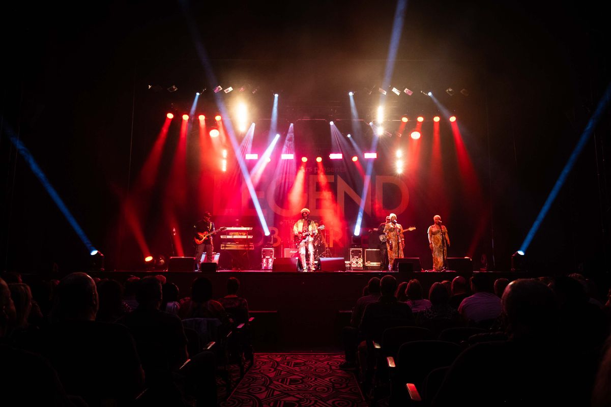 A vibrant live concert scene featuring a singer and musicians on stage under red and blue lights. An audience watches, creating an energetic atmosphere.