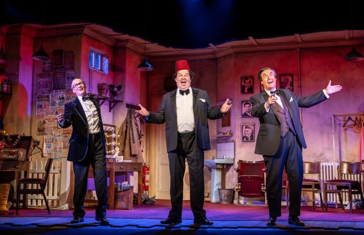 Three men stood together singing in a dressing room backstage type setting, one is wearing a fez and they are all dressed smartly