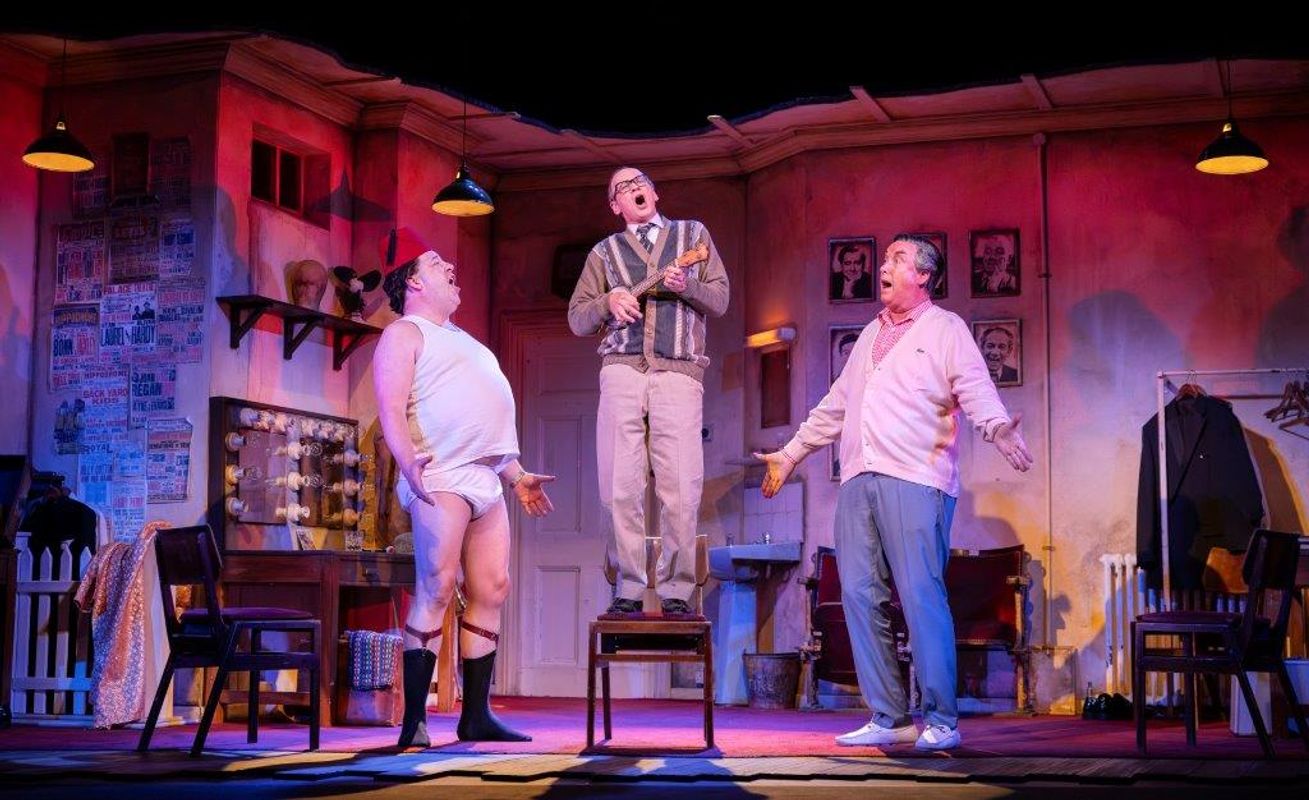 Three actors perform in a lively theater scene. One stands wearing a fez, another stands on a chair singing, with glasses, and the third smiles in pastel clothing. The room is adorned with posters and framed photos. The setting is warmly lit, creating a comedic and vibrant atmosphere.
