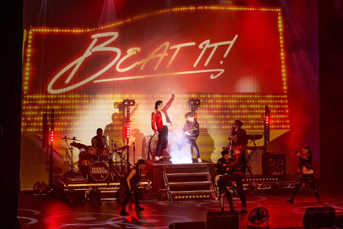 Stage performance of "Beat It" with vibrant lighting, dancers in dynamic poses, and musicians. The scene is energetic and lively, capturing the essence of a live show.