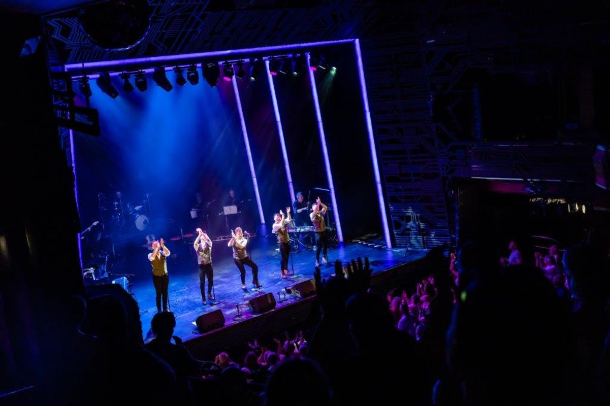 A band performs energetically on a vibrant stage with colorful lighting and vertical neon accents. An enthusiastic audience claps and cheers.