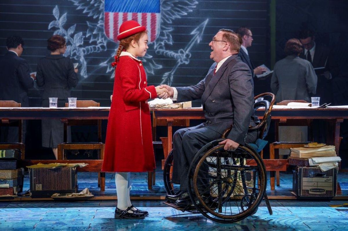 There is a young girl in a red coat and hat and a gentleman in a wheelchair. they are shaking hands