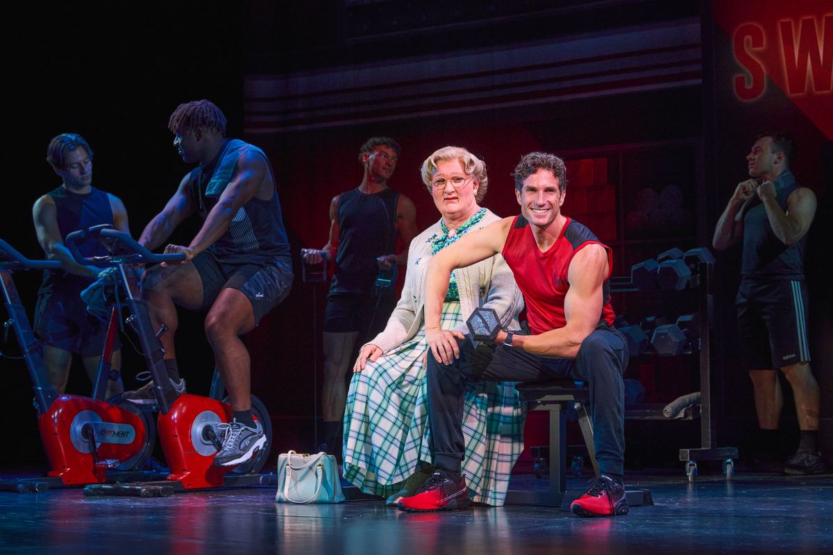 Mrs Doubtfire sits in the middle of a gym next to a muscly man lifting weights.