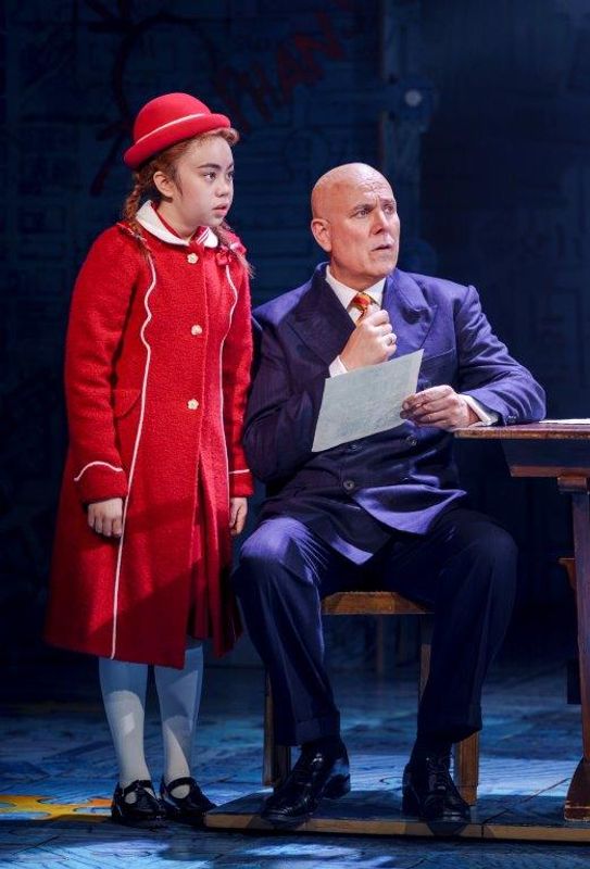 There is a young girl in a red coat and hat. A gentleman is sat down at a desk and she is stood beside him