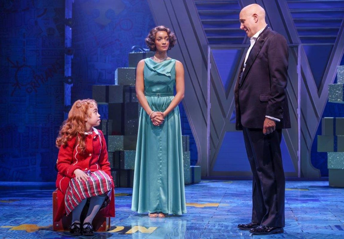There is a young girl in a red coat sitting down, She is looking up at a gentleman in a suit. There is a lady in a blue floor length dress stood watching them