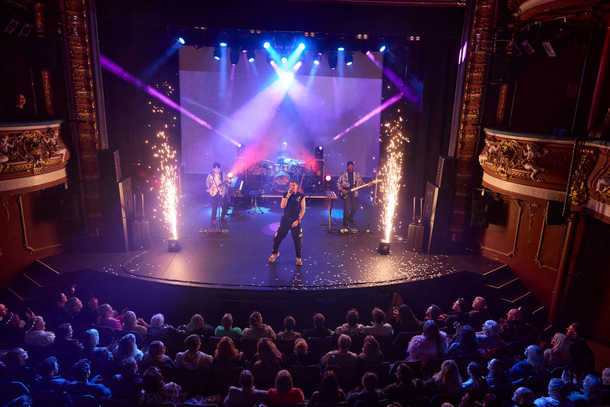 A vibrant concert scene. A singer and band perform on stage, surrounded by purple and blue lights with fireworks. Audience watches intently.
