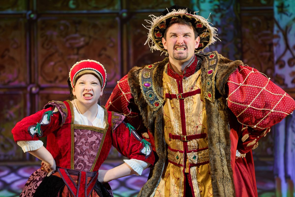 Two people on stage, they are all wearing 14th century clothing. They both have their hands on their waists with angry expressions on their faces