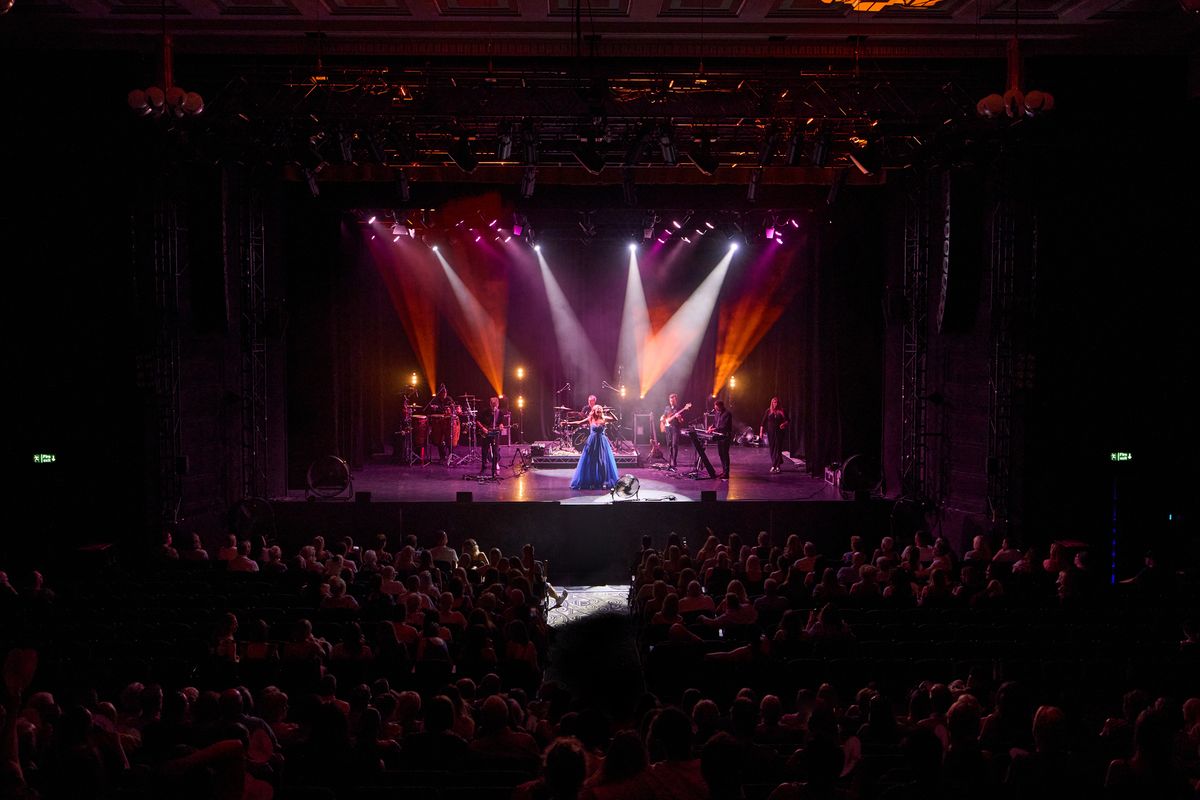 A performer in a blue outfit stands on a brightly lit stage with various colored lights. A band or group of musicians plays in the background. The audience is seated in a theater, watching the performance.