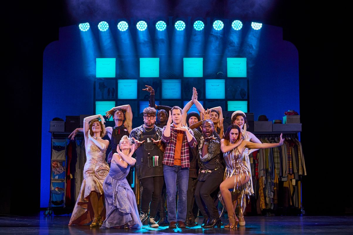 Daniel stands centre stage in a chequered shirt surrounded by an ensemble all doing Vogue dance moves.