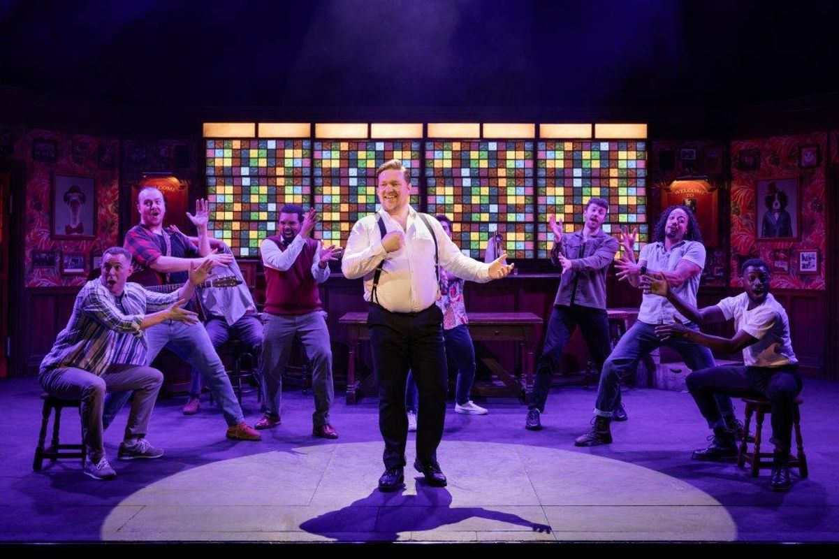 One person is stood in the centre with a spot light on them, there is a group of people surrounding them with their hands out