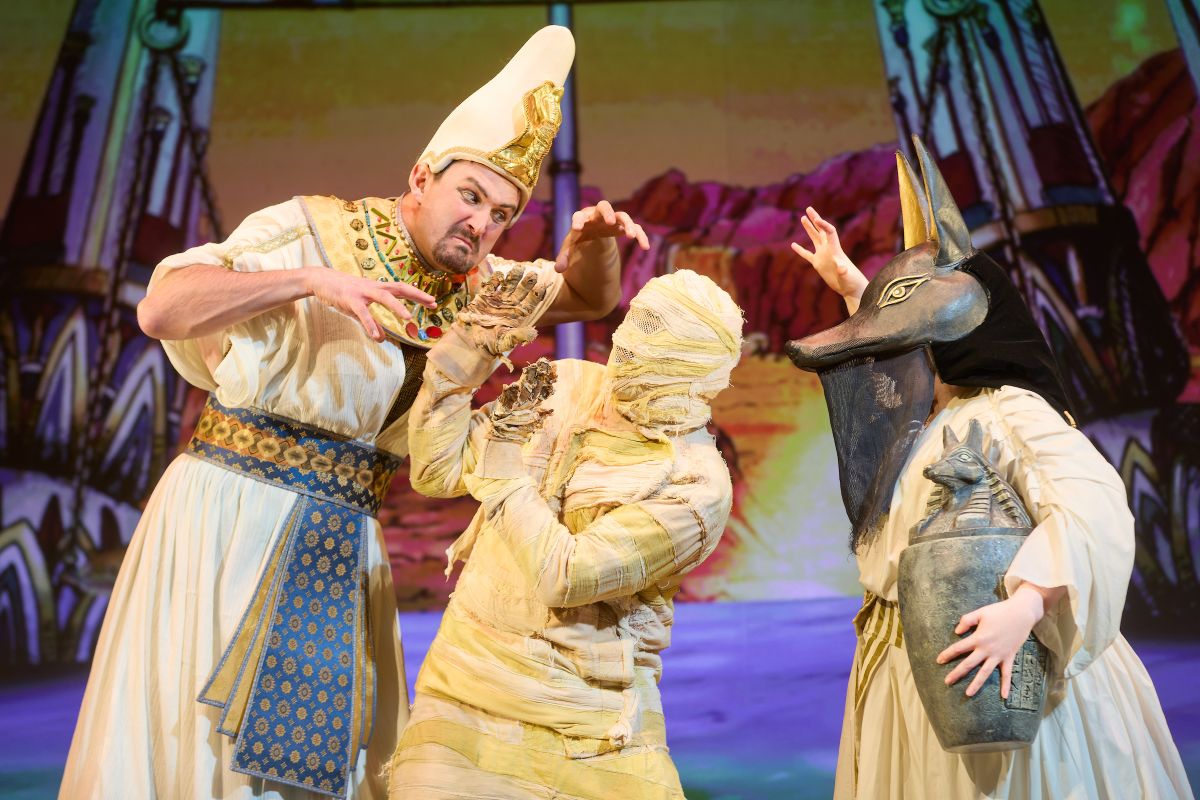 Three people on stage, one is dressed as an ancient Egyptian mummy, they are positioned in the middle. There are  people either side of the ancient mummy and they are dressed in ancient Egyptian clothing