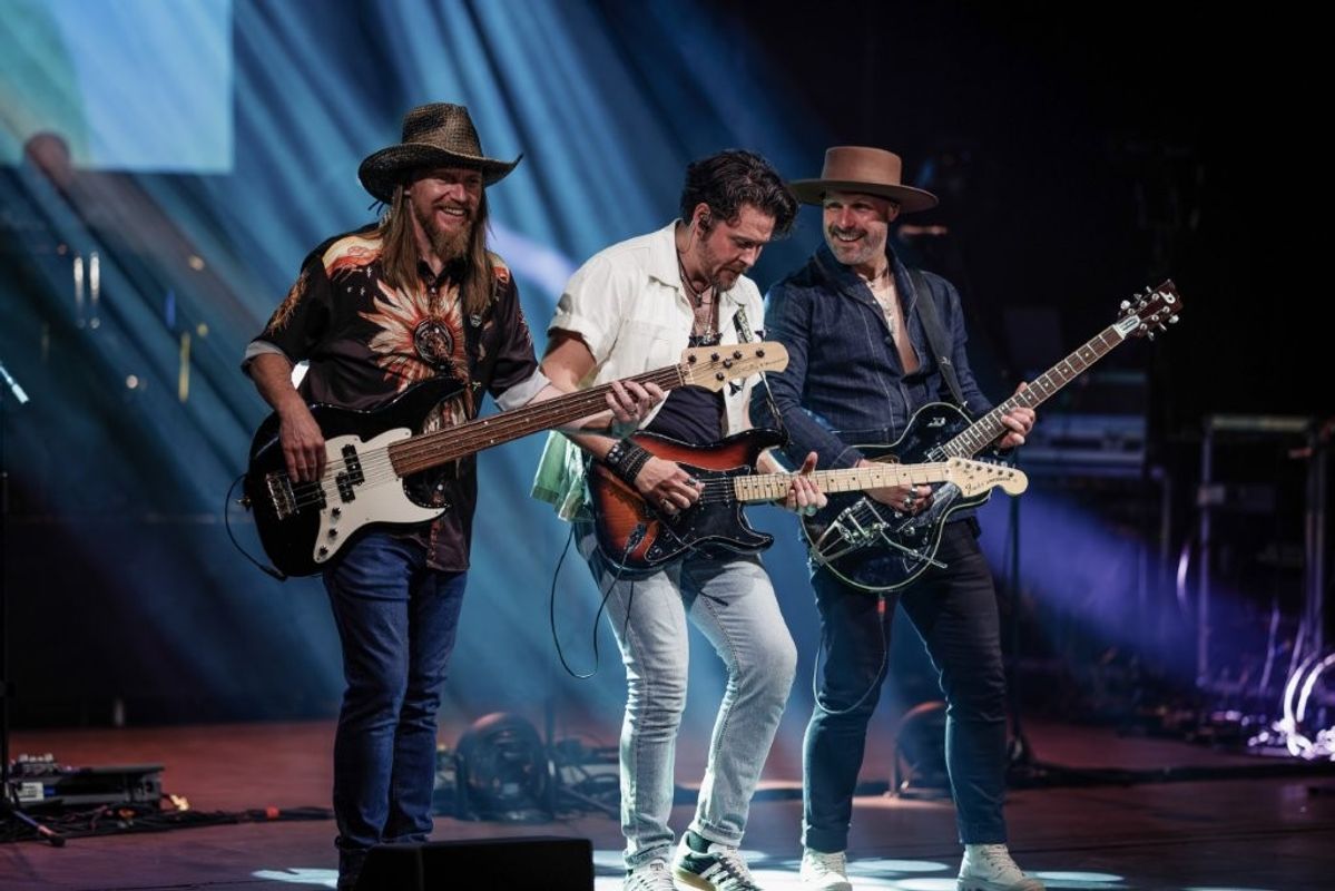 Three musicians perform energetically on stage, each playing a guitar. Stage lights create a dynamic atmosphere, highlighting their expressions of joy.