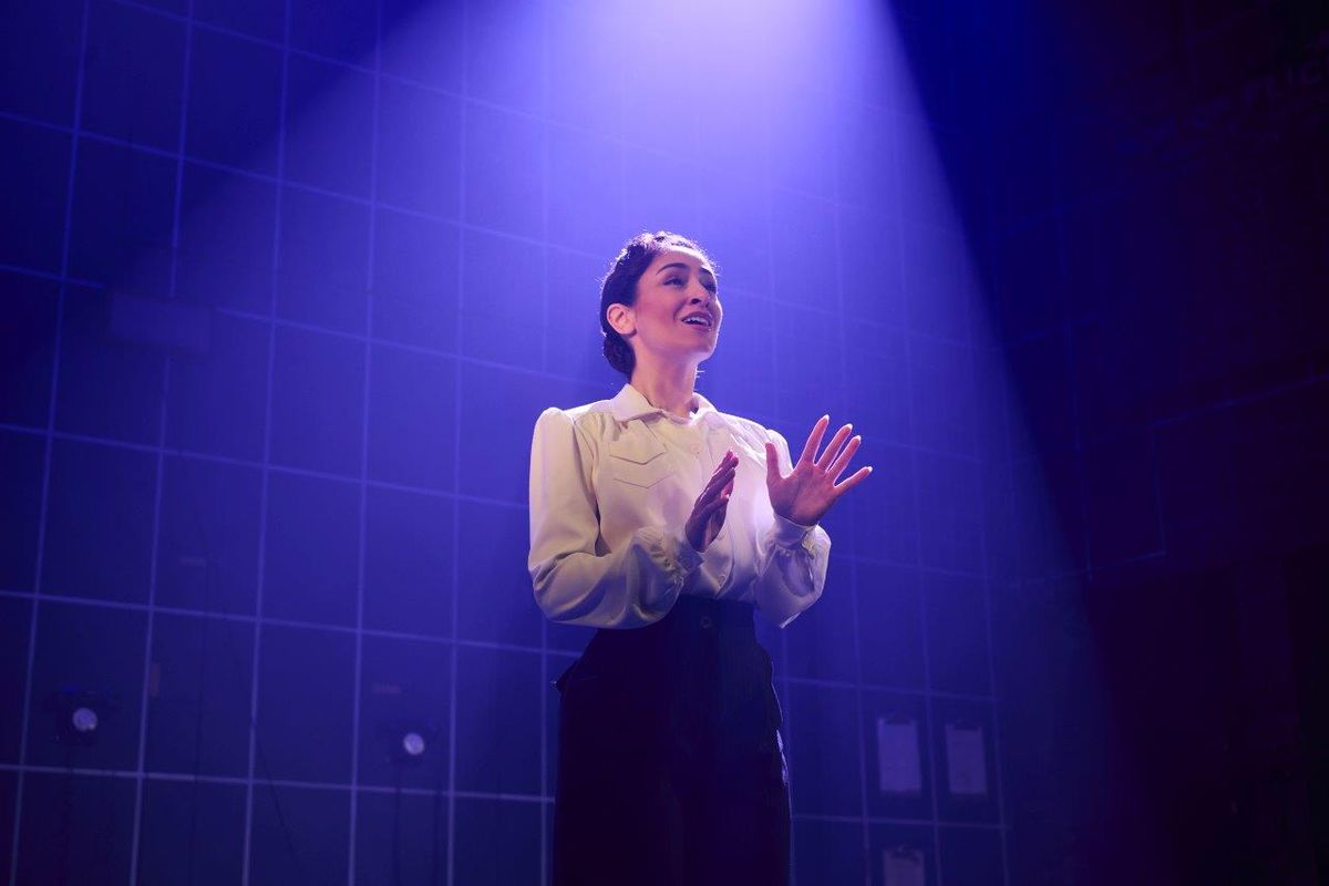 A woman in a white shirt on stage, a spot light is shining down on her