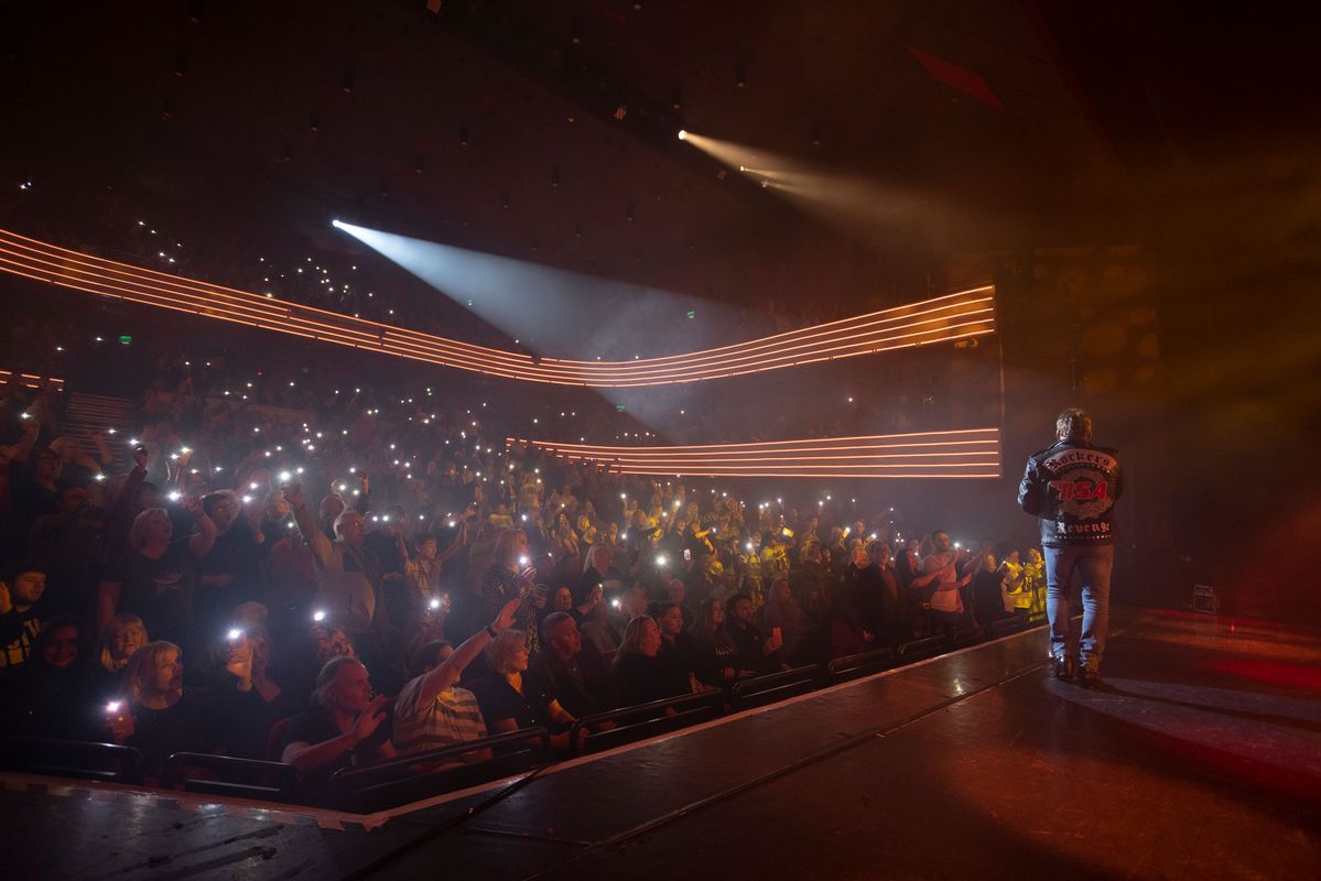 A man performing on stage to an audience. There are colourful lights. Behind him are backup singers and a band playing