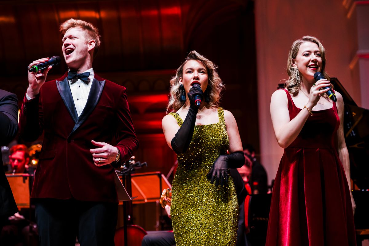 Three singers wearing velvet outfits stand on stage.