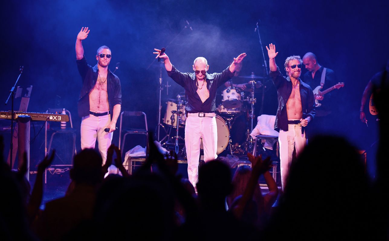 Three people walk towards the front of the stage wearing white trousers and  unbuttoned black shirts.