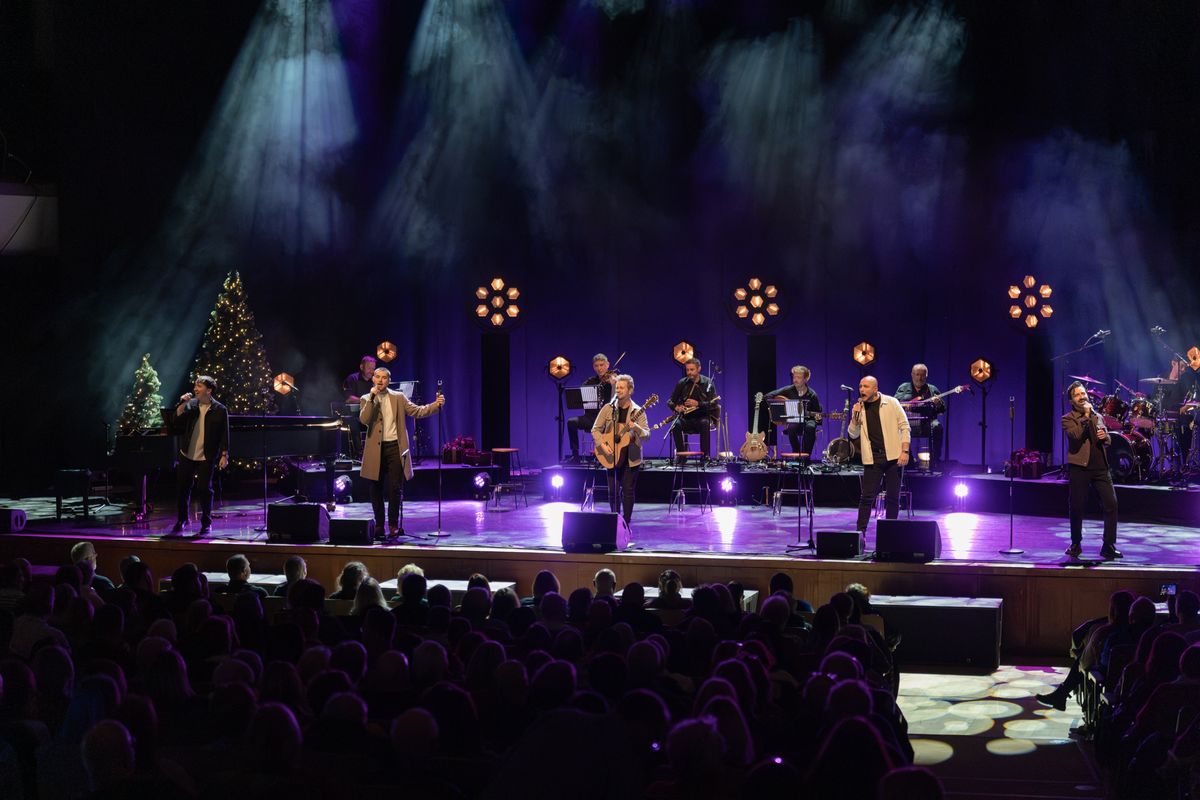 The five band members are performing on stage, one member in the middle is singing with a guitar, the other four members stood either side are singing into microphones. There are people playing instruments behind them. There is purple lighting being shone onto the stage