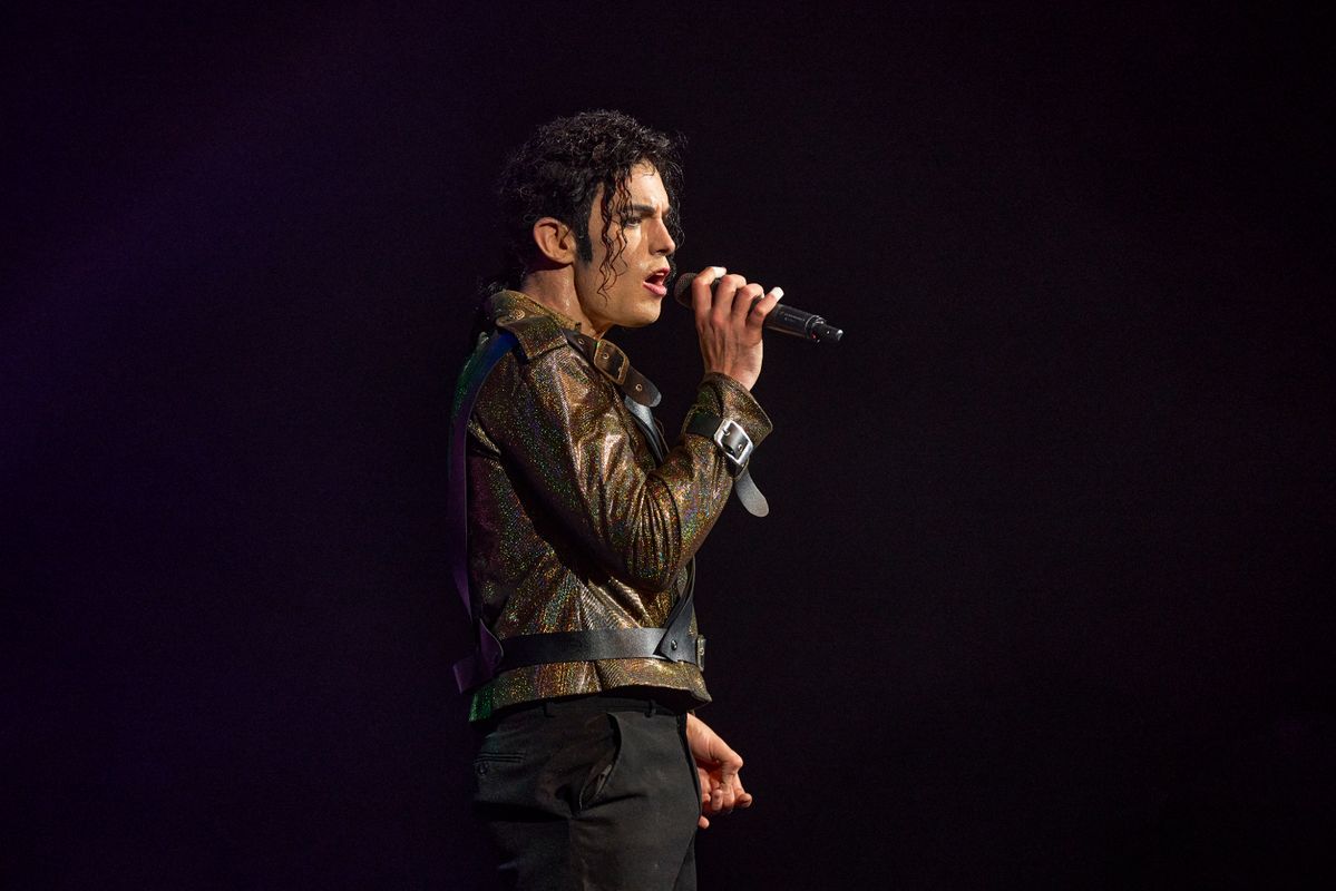 A person sings passionately into a microphone on stage, wearing a sparkly jacket and styled curly hair, with a focused expression, in dim lighting.