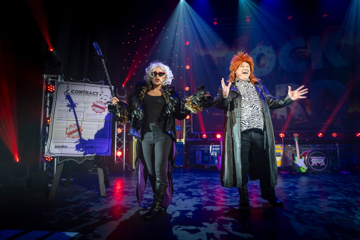 Two people stand in a spotlight on stage wearing wacky outfits.