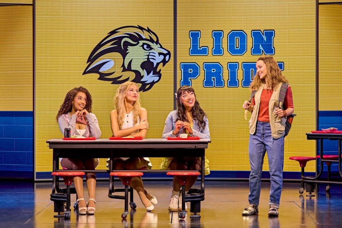 Four Mean Girls cast members sit at a red cafeteria table on stage. Behind them, a "Lion Pride" logo is displayed on a yellow wall. They appear engaged and cheerful.