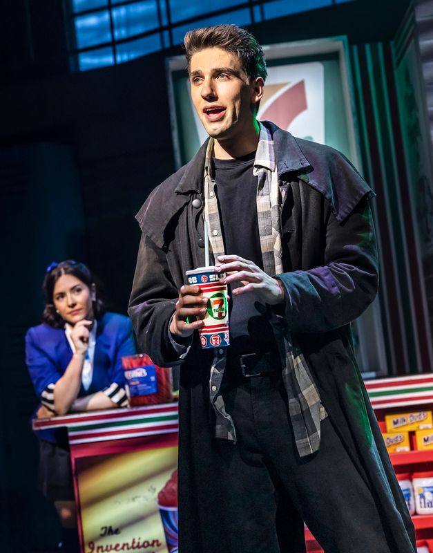 A man in a dark coat sings passionately while holding a 7-Eleven cup on stage. A woman in a blue uniform watches attentively from a counter. Stage lighting creates a dramatic ambiance.