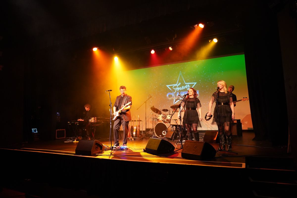 A person plays guitar on stage whilst backed by two singers.