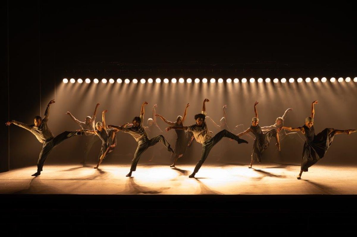 Group of dancers on stage wearing blue and grey outfits, a light casts a shadow over them. They are all posing the same with a leg up and their arms in the air