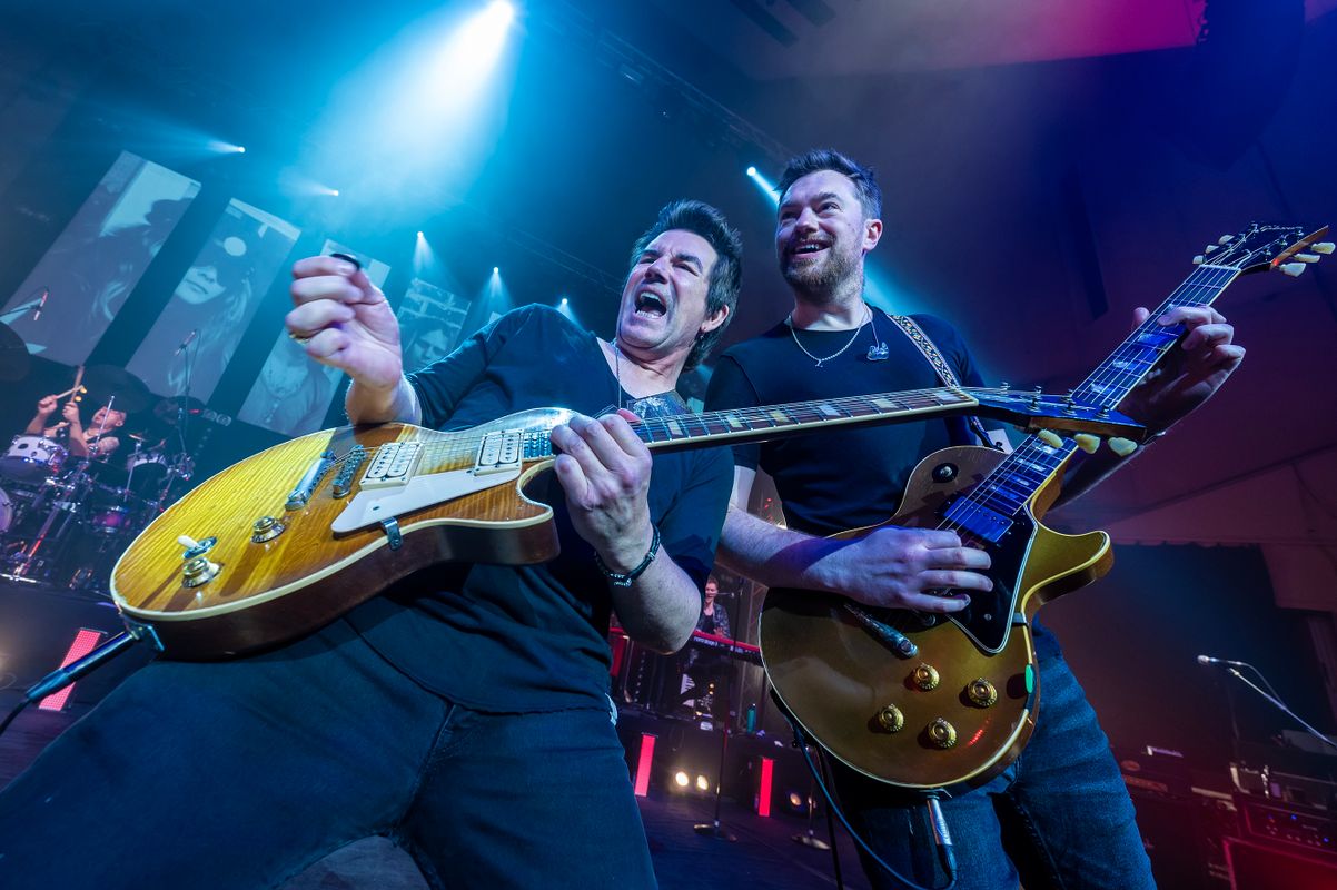 Two people stand shoulder to shoulder playing guitar.