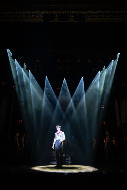 An actor with a red neckerchief and bloodied face stands centre stage surrounded by spotlights.
