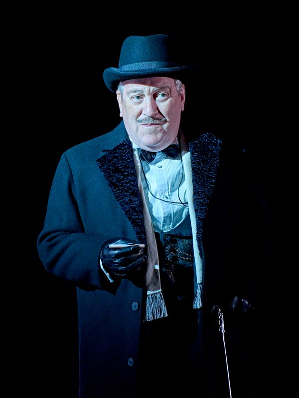 A person dressed in a black coat, black bowler hat and leather gloves.