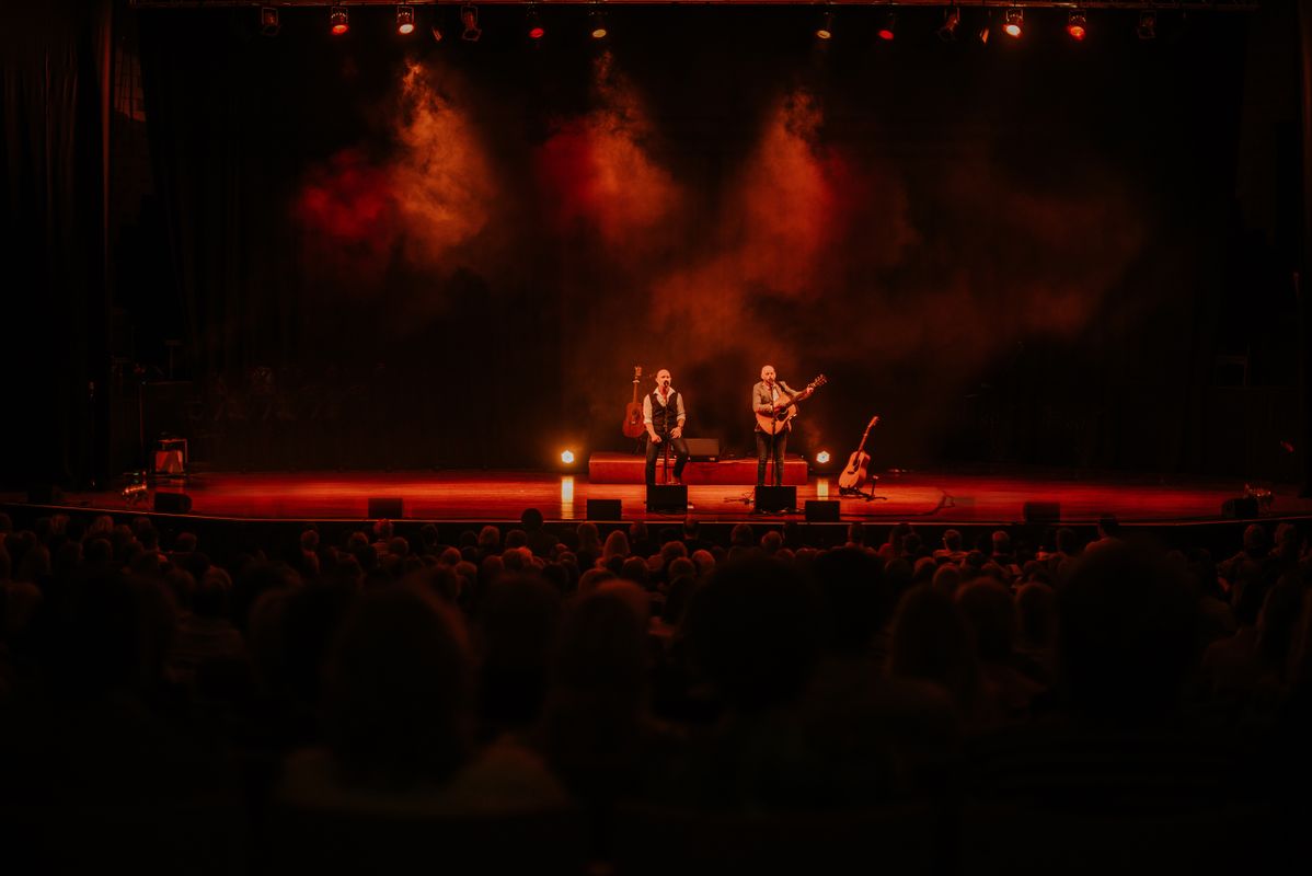One person is singing into a microphone and the other person singing into a microphone and playing the guitar