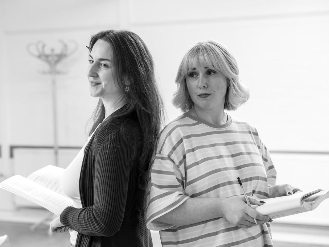 Two woman are stood back to back with each other. They are both holding scripts