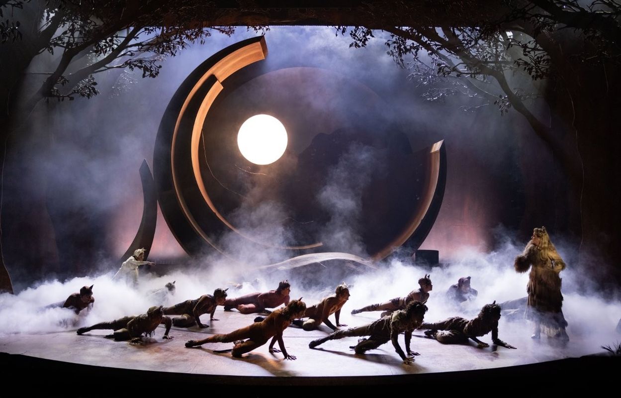 A theatrical scene from "Cats" features performers in animal costumes, resembling felines, crouched on stage surrounded by mist. A large, circular set piece at the back resembles a rising sun, with tree silhouettes framing the scene.