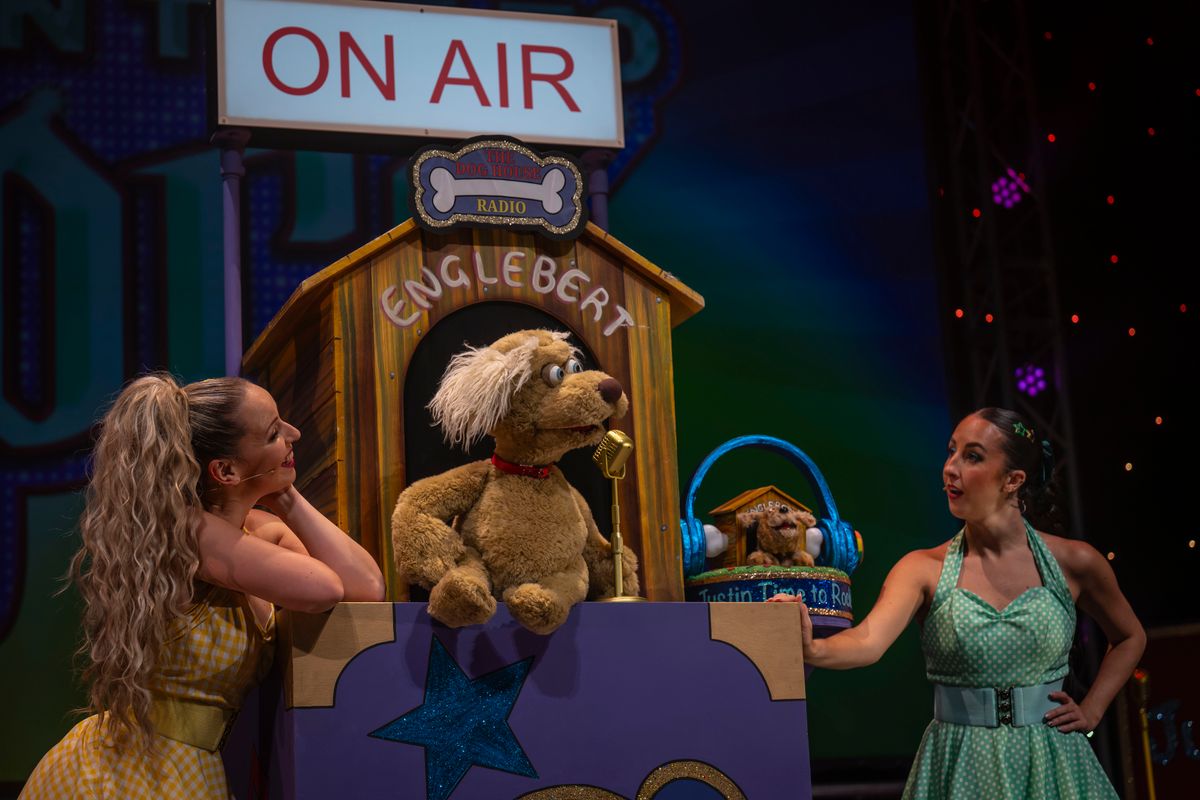 A puppet dog is sat in front of a dog house singing into a microphone with two ensemble members either side.