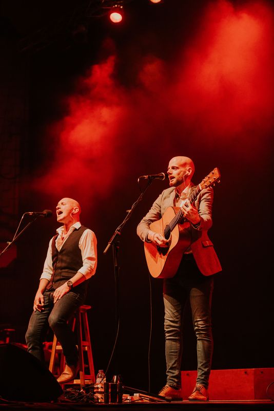 One person is singing into a microphone and the other person singing into a microphone and playing the guitar