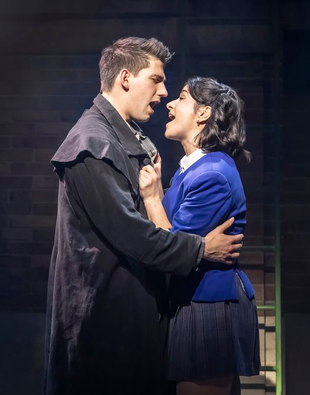 A young man in a dark coat and a woman in a blue blazer passionately sing to each other on stage, conveying strong emotion and intensity.